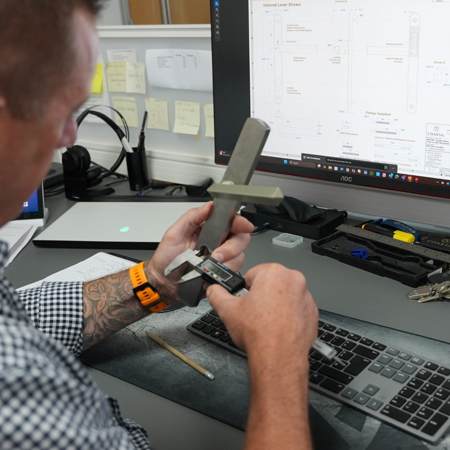 Person using a caliper at a desk with computer monitors and office supplies.